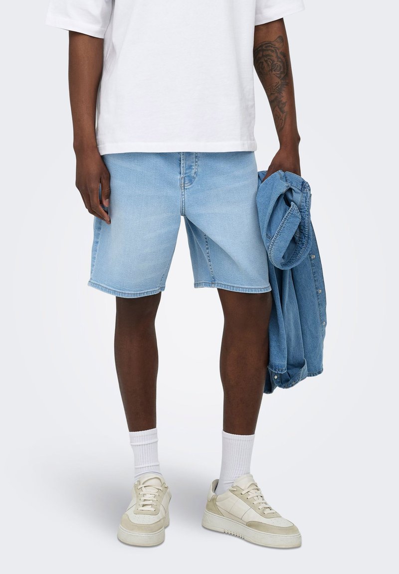 Personne portant un short en jean bleu clair, des chaussettes blanches, des baskets blanches, un t-shirt blanc et tenant une veste en jean bleue dans une main.