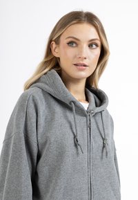 Szürke, kapucnis, cipzáros kapucnis pulóver, puha anyagból készült. Húzózsinórral, elülső cipzárral és laza szabással rendelkezik. Nincsenek minták vagy díszítések.