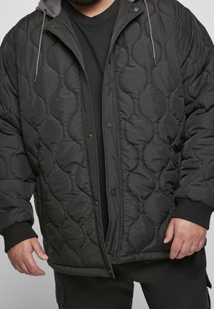 Homme portant une veste matelassée noire avec boutons-pression, poignets côtelés et capuche avec cordons sur une chemise noire.