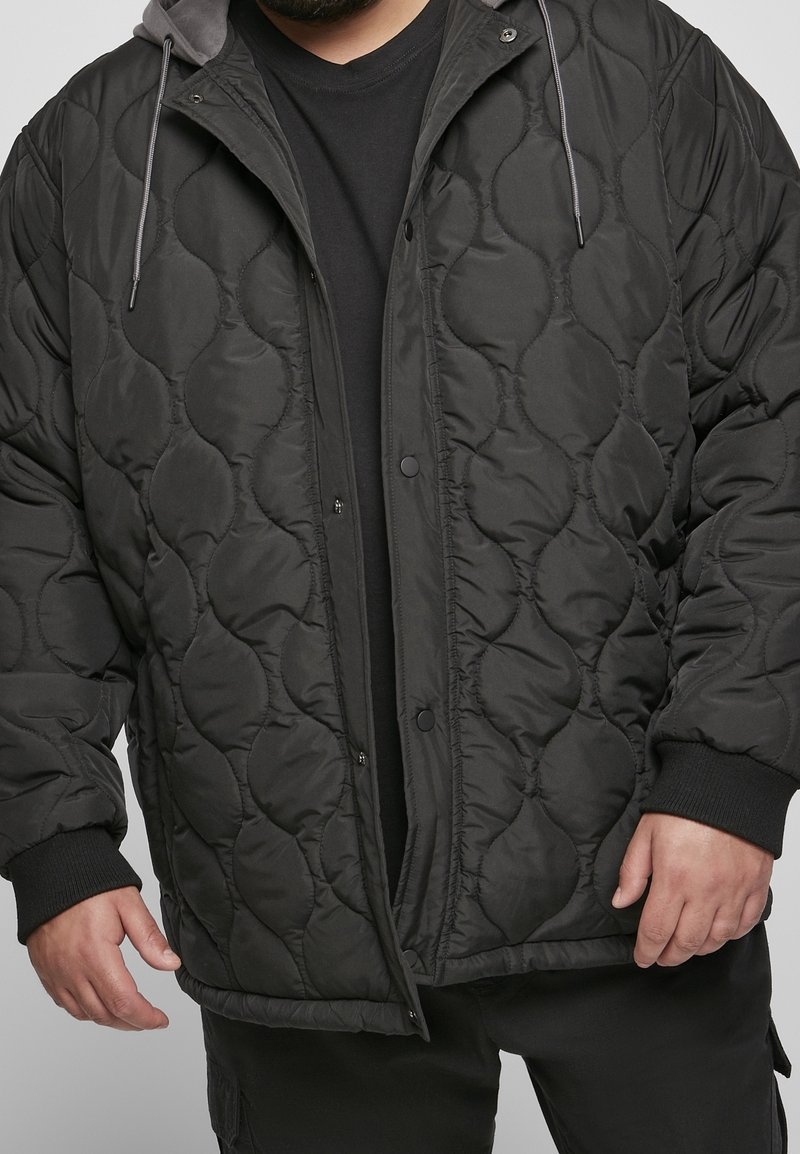 Homme portant une veste matelassée noire avec boutons-pression, poignets côtelés et capuche avec cordons sur une chemise noire.