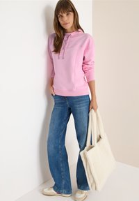 Roze sweatshirt met een capuchon, koordjes en geribbelde manchetten, gecombineerd met blauwe flare jeans en een witte fluffy schoudertas.