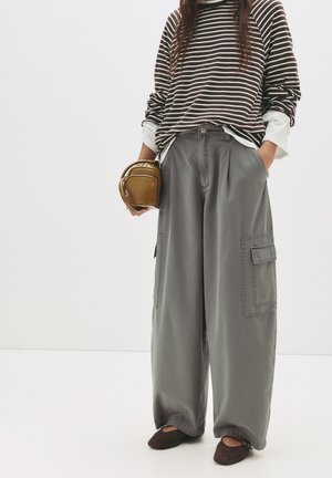 Grijze wijde cargo-broek met zijzakken, gecombineerd met een gestreepte long-sleeve shirt en bruine instappers, met een bruine tas in de hand.