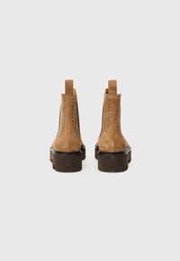 Bottes chelsea en daim marron avec une semelle robuste, présentant des détails cousus et des languettes à l'arrière. Fini texturé et forme arrondie.