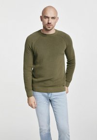 Oliwkowy, prążkowany sweter z okrągłym dekoltem i długimi rękawami. Noszony z jasnoniebieskimi dżinsami, z subtelnym detalem logo.