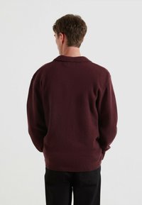 Bordowy wełniany sweter o luźnym kroju, z kołnierzykiem i ściągaczami na rękawach, pokazany z tyłu na jednolitym tle.