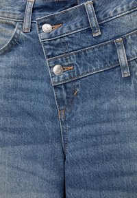 Jeans de mezclilla azul claro con dos botones plateados, costuras naranjas en contraste y una textura suave; con un diseño clásico de cinco bolsillos.