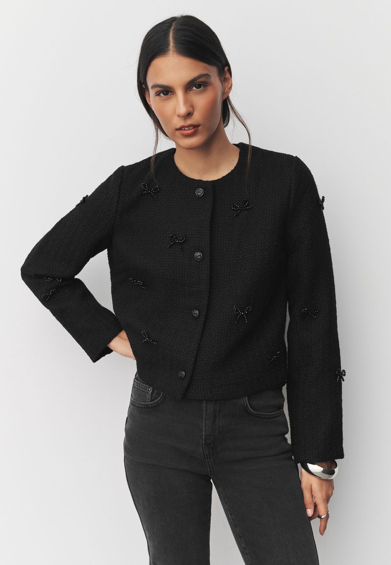 Jeune femme aux cheveux noirs et raides porte une veste noire boutonnée avec de petites décorations en forme de nœuds et un jean foncé taille haute.