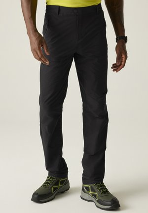 Hombre que lleva pantalones de exterior negros con bolsillos con cremallera y zapatillas de senderismo verdes y negras, de pie contra un fondo claro.