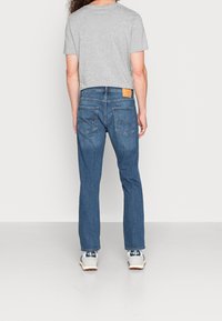 Světle šedé tričko s krátkým rukávem v kombinaci s tmavě modrými džínami. Džíny mají koženou nášivku na zadní straně a klasický pětikapsový design.