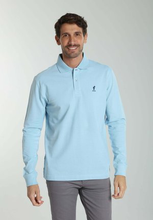 Homme souriant portant un polo bleu clair à manches longues et un pantalon gris, debout devant un fond clair uni.