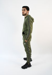Sudadera y pantalones jogger verde olivo de tela suave, con rayas laterales negras que llevan texto blanco. Zapatillas deportivas negras.