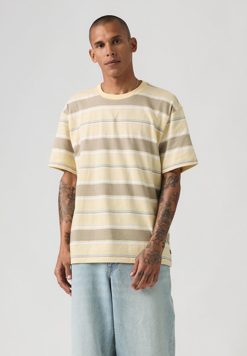 Homme aux cheveux courts et tatouages portant une chemise beige à manches courtes rayée et un jean bleu clair, debout devant un fond uni clair.