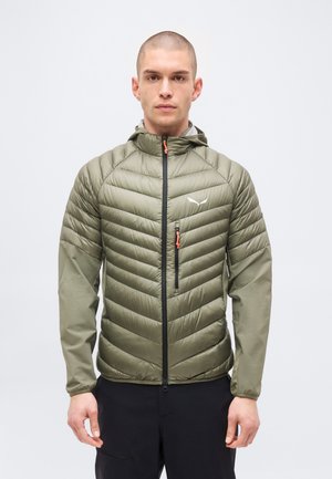 Uomo con testa rasata che indossa una giacca imbottita verde oliva con cappuccio e cerniera nera, in piedi davanti a uno sfondo chiaro e uniforme.