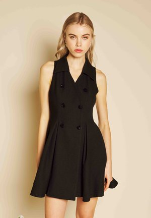 Junge Frau mit blonden Haaren, die ein ärmelloses schwarzes, zweireihiges Kleid vor einem beigen Hintergrund trägt.