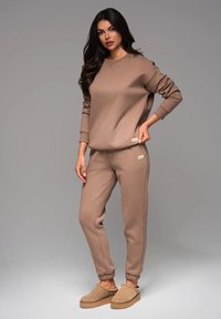 Braunes Sweat-Outfit aus weichem Stoff; lockeres Sweatshirt, schmal zulaufende Jogginghosen mit gerippten Bündchen und passende beigere Hausschuhe.