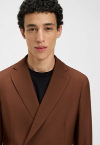Brauner, taillierter Blazer mit Spitzrevers und dezenten Steppdetails, getragen über einem schwarzen Hemd. Der Stoff wirkt glatt und hat eine strukturierte Passform.