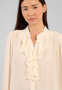 Blusa crema con fronte arricciato, chiusura a bottoni e maniche a sbuffo arricciate. Realizzata in tessuto leggero con una trama liscia.