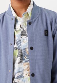 Giacca a maglia azzurro chiaro con texture a coste, chiusura con bottoni e una piccola toppa nera con logo, indossata sopra una camicia floreale a fantasia.
