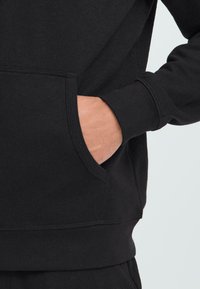 Mano inserita nella tasca anteriore di un hoodie nero a maniche lunghe, che mostra il polso e parte dell'avambraccio su uno sfondo chiaro.
