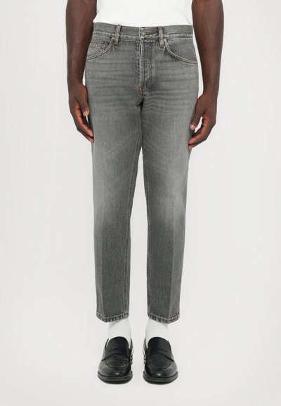 Man wearing gray straight-leg jeans, white socks, black loafers, and a white shirt standing against a plain background.