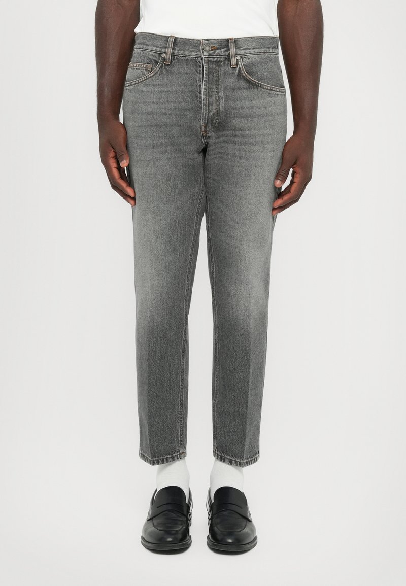 Man wearing gray straight-leg jeans, white socks, black loafers, and a white shirt standing against a plain background.