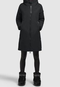 Abrigo de invierno largo y negro con cremallera frontal, manos en los bolsillos, combinado con leggins negros brillantes y botas de invierno negras sobre fondo liso.