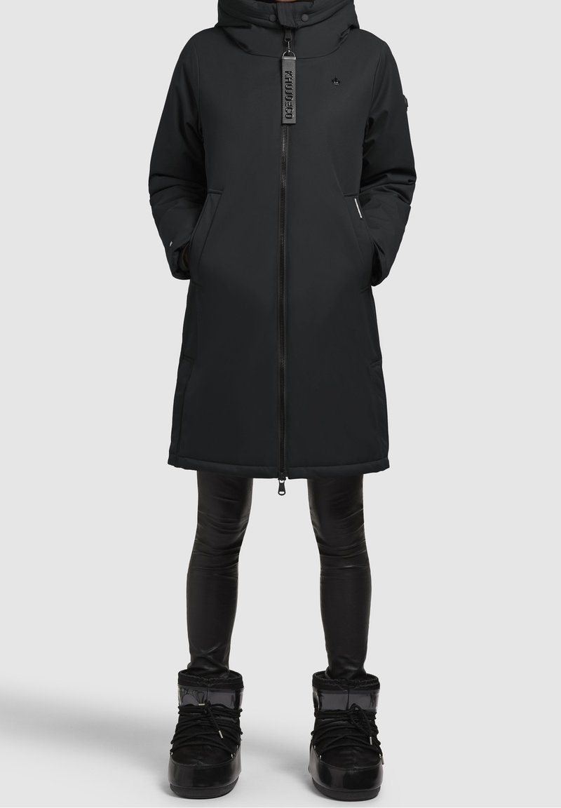 Abrigo de invierno largo y negro con cremallera frontal, manos en los bolsillos, combinado con leggins negros brillantes y botas de invierno negras sobre fondo liso.