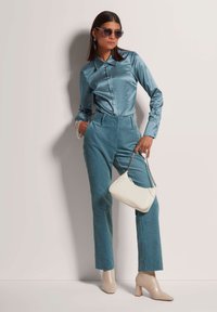 Türkise Satinbluse kombiniert mit ribbed, petrolfarbenen Hosen. Das Model hält eine weiße Handtasche mit einer silbernen Kette. Beigefarbene spitze Absatzschuhe runden den Look ab.