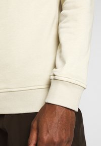 Lys beige hettegenser med ribbestrikkede ermer og kant, laget av mykt stoff. Plagget har en jevn tekstur med minimale detaljer.