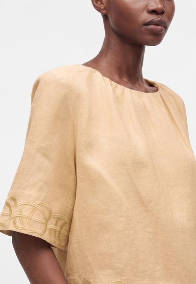 Femme portant un haut en lin beige avec des motifs circulaires brodés sur les manches et près de l'ourlet, montrée du cou jusqu'au milieu du torse.