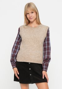 Beige pletený svetr s ribovanou strukturou, přehozený přes kostkovanou košili s dlouhým rukávem v tmavě červené a modré, doplněný černou manšestrovou sukní.