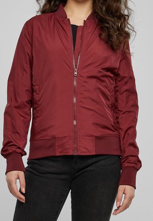 Femme portant une veste bomber bordeaux zippée sur un t-shirt noir et un pantalon noir, debout sur un fond gris clair uni.