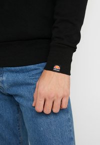 Sudadera negra con puños acanalados y un pequeño logo de ellesse de color. Llevada con unos vaqueros de mezclilla azul que muestran una tela texturizada.