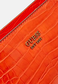 Pomarańczowa skórzana tekstura z wzorem krokodyla oraz złotym logo "GUESS EST 1981" na akcesorium modowym.