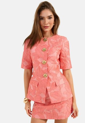 Young woman wearing coral floral textured short-sleeve blazer with gold flower buttons and matching short skirt, standing against white background.
