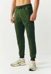 Grüne Jogginghose mit weicher Textur, elastischem Bund, Reißverschlusstaschen, schmal zulaufenden Beinen und Rippbündchen, kombiniert mit beigen Sportschuhen.