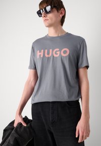 Pilkas medvilninis marškinėlis su ryškiai rožiniu užrašu "HUGO", derinamas su juodais džinsais. Modelis laiko juodą švarką. Juodi akiniai nuo saulės.