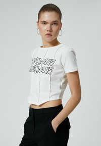 Valkoinen lyhythihainen cropped-t-paita, jossa on mustaa graffitotyylistä tekstiä. Kangas näyttää sileältä, ja saumat ovat selkeästi näkyvissä.