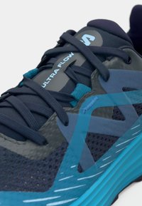 Chaussure de course pour hommes en bleu marine et bleu sarcelle. Présente une tige en mesh avec des accents texturés et une semelle en caoutchouc. Marque "ULTRA FLOW" sur la languette.