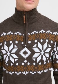 Maglione marrone lavorato a maglia con colletto alto, dotato di chiusura con zip e modelli di fiocchi di neve bianchi, arancioni e gialli sulla parte anteriore.