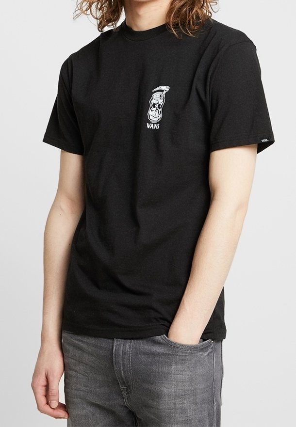 Personne portant un t-shirt Vans noir avec un petit graphique blanc d’un crâne et d'une bombe de peinture sur la poitrine, assorti à un jean gris.