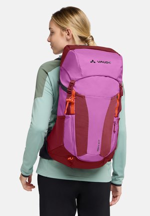 Donna con capelli biondi che indossa una giacca verde smeraldo e pantaloni neri, con un grande zaino da trekking viola e rosso con l'etichetta "Vaude".