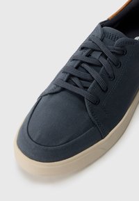 Sapatilha de lona azul-escura com bico arredondado, atacadores sintéticos e sola de borracha bege; possui costura e elementos de design minimalistas.