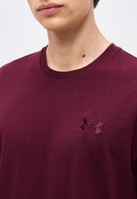 Νέο άτομο φορεί ένα σκούρο κόκκινο T-shirt Under Armour, εμφανίζοντας τον λαιμό με ορατή ελιά και μέρη από τα χείλη και το πηγούνι σε μονόχρωμο φόντο.