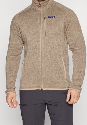 Hombre con chaqueta de forro polar beige de Patagonia con cremallera, dos bolsillos frontales y un bolsillo con cremallera en el pecho, combinado con pantalones oscuros.