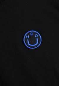 Black cotton fabric featuring a round blue embroidered logo with the letters "HGO" and a smiley face design. Textured, with a smooth finish.