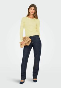 Ljusgul ribbad långärmad topp med en scallop-edged nederkant, ihop med mörkblå raka jeans. Håller en vävd väska och har på sig blå klackar.