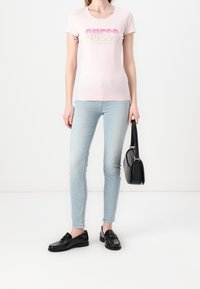 Kobieta ubrana w jasnoróżową koszulkę "GUESS Los Angeles", jasnoniebieskie dżinsy, czarne mokasyny, trzymająca czarną torebkę przy boku.