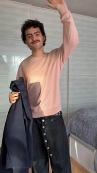 Jeune homme en sweat-shirt rose et pantalon noir boutonné souriant, tenant une veste sombre et faisant signe de la main à l'intérieur près d'un lit avec une couverture grise.