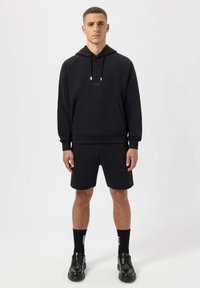 Black hoodie with adjustable drawstrings, front pocket, and logo. Paired with black shorts, black crew socks, and black lace-up shoes.